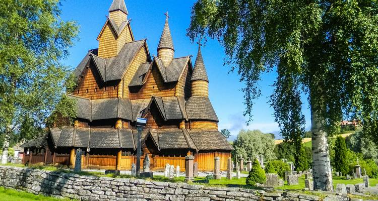 Historische houten staafkerk met meerdere steile daken gelegen naast bomen en grafstenen.
