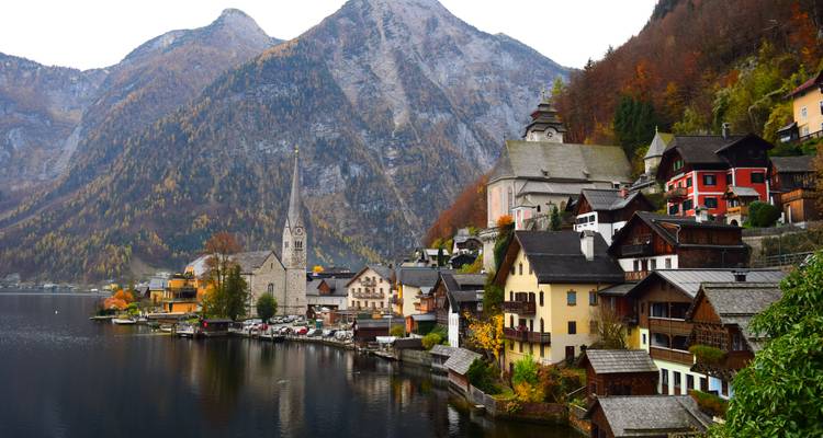 Ein herbstlicher Blick auf Hallstatts Kirchturm und die dicht gedrängten Häuser am ruhigen See unterhalb schroffer Berge, die in herbstlichen Farben erstrahlen.