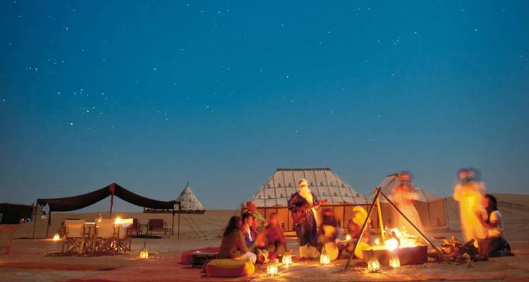 Campamento en el desierto por la noche con personas alrededor de una fogata brillante, tiendas tradicionales y cielo estrellado arriba.