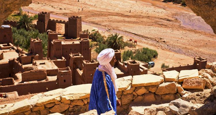 Hombre bereber con túnica azul contempla un antiguo pueblo kasbah de tierra y el valle desértico que se extiende abajo.