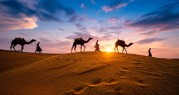 Siluetas de camellos y nómadas coronando una duna contra un cielo de atardecer vívido.