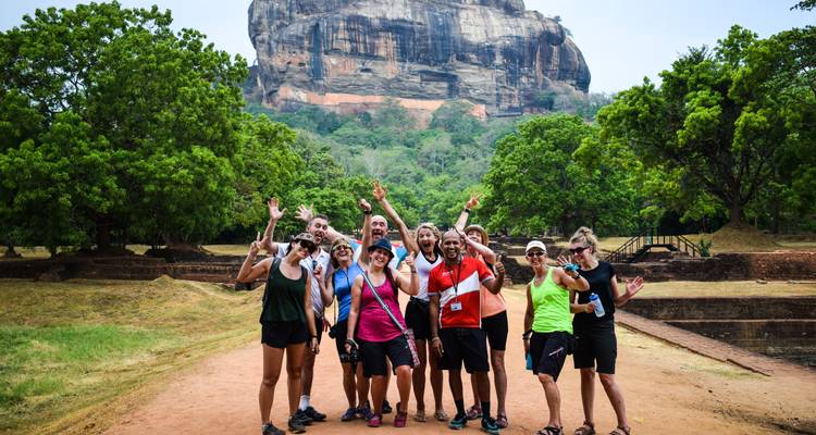 Un groupe de touristes jubilant pose sur un sentier avec l'emblématique forteresse rocheuse de Sigiriya qui se dresse derrière des arbres luxuriants.
