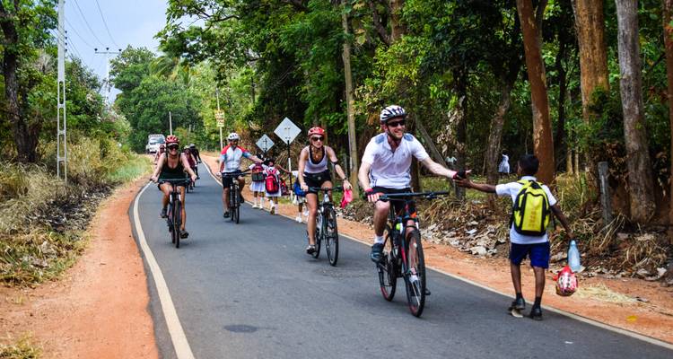 Des cyclistes roulent sur une route de campagne bordée de grands arbres, échangeant un tape-m'en-cinq joyeux avec un enfant du coin portant un cartable.