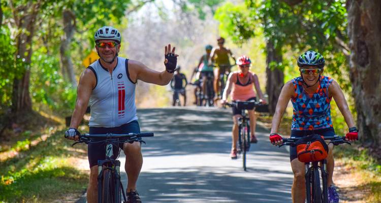 Une file de cyclistes fait signe depuis un chemin de campagne ombragé encadré par un feuillage vert vif et une lumière tachetée du soleil.