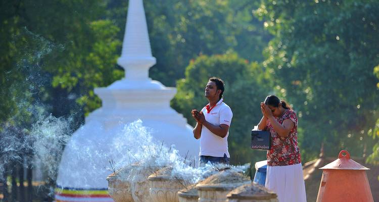 Deux fidèles se tiennent devant des brûle-encens fumants face à un stupa blanc au milieu d'une végétation luxuriante.