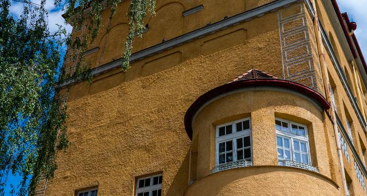 Nahaufnahme eines senfgelben Stuckgebäudes mit gewölbten Fenstern und dekorativen Verzierungen unter einem hellen Himmel.