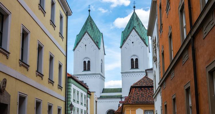 Eine schmale Altstadtgasse rahmt zwei weiße Kirchtürme mit Spitzdächern ein, die grüne Dächer haben und sich gegen einen blauen Himmel abheben.