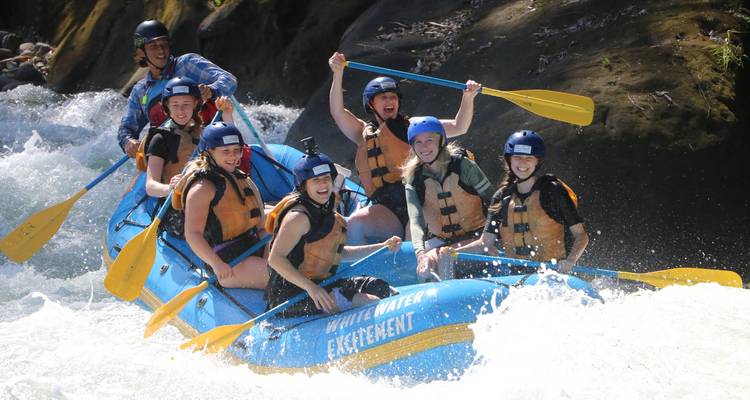 Excited group of paddlers in blue helmets navigating rapids in a bright blue raft, water splashing dramatically.