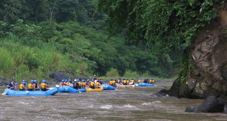 Multiple rafts filled with adventurers float down a wide tropical river framed by steep green canyon walls.