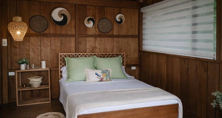 Cozy wooden cabin bedroom with green pillows, woven wall decor and filtered daylight.
