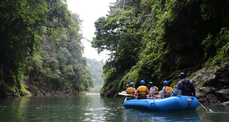Raft with life-jacketed travelers gliding through a serene green gorge surrounded by lush rainforest.