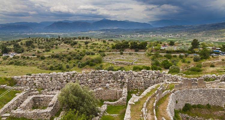 Stone ruins and terrace walls of Mycenae looking over a fertile plain under dramatic clouds.