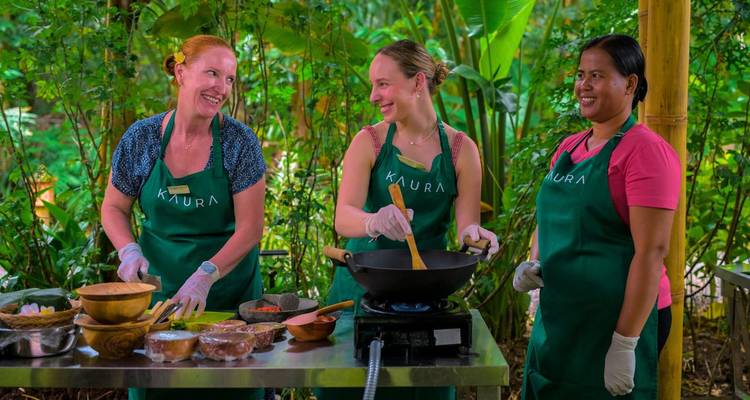 Drie vrouwen in schorten delen een lach tijdens een Balinese kookles buiten.