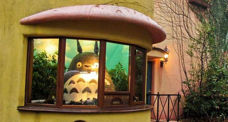 Totoro figuur die door een gebogen museumraam van een grillig geel gebouw kijkt.