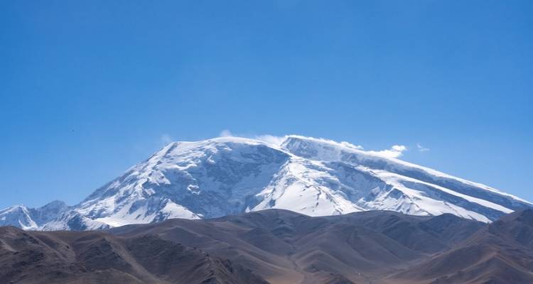 Sommet montagneux enneigé s'élevant au-dessus de contreforts arides et bruns sous un ciel bleu dégagé.
