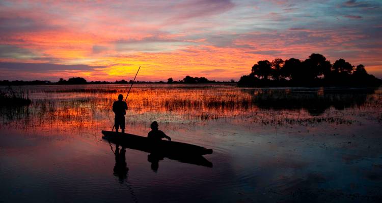 Des percheurs en silhouette naviguent en mokoro à travers des zones humides brillantes sous un coucher de soleil orange éclatant.