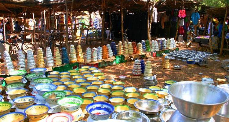 Colorful ceramic bowls fill a bustling outdoor market stall under rustic thatched roofs.