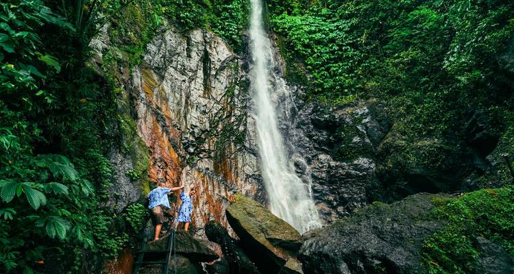 Twee bezoekers bewonderen een hoge regenwoudwaterval die over natte rotsen naar beneden valt in een poel eronder.