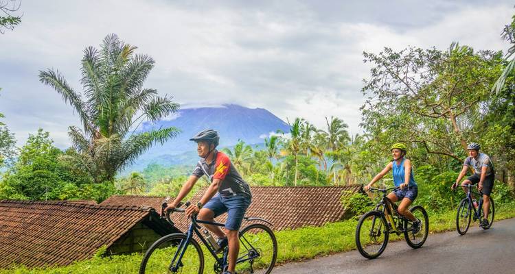 Fietsers rijden over een landelijke weg met een vulkaan en palmbomen op de achtergrond onder bewolkte luchten.