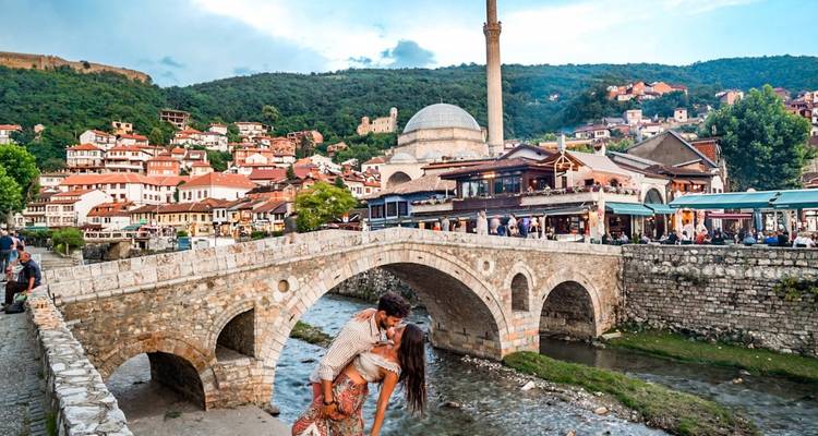 Un couple s'embrasse sur le pont de pierre historique de Prizren avec la ville animée à flanc de colline en arrière-plan.