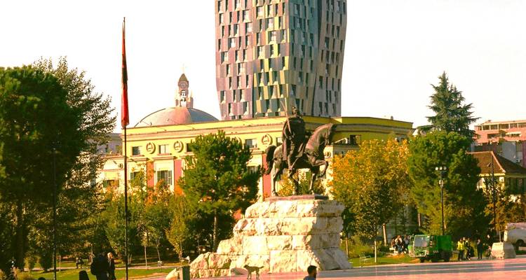 Statue de Skanderbeg sur la place centrale de Tirana avec des bâtiments modernes et historiques en arrière-plan.
