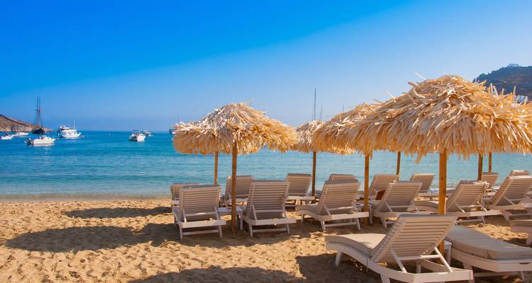 Zandstrand met rijen ligstoelen onder rieten parasols met uitzicht op turquoise water en geankerde jachten