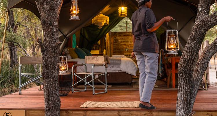 A staff member carries lanterns on a wooden platform outside a canvas safari tent at dusk.