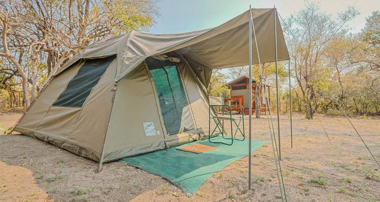 A khaki safari tent with an awning is pitched on sandy soil under leafy trees.