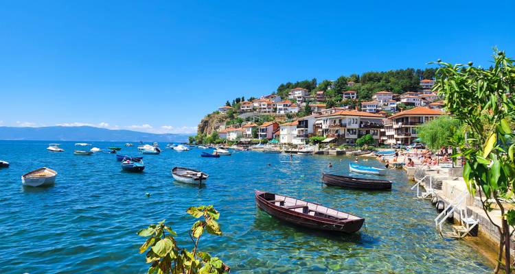 Lago Ohrid azul cristalino salpicado de pequeños botes y pueblo en la ladera bajo cielo brillante