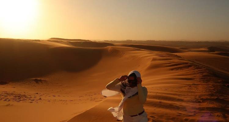 Traveler in headscarf standing atop golden desert dunes at sunrise with warm glow.