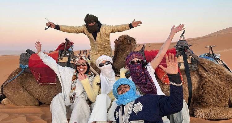 Group of friends in traditional scarves posing happily with camels on desert sands during twilight.