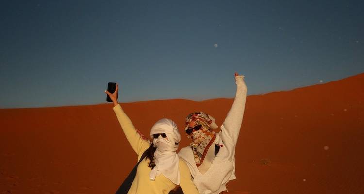 Two travelers celebrating with raised arms against smooth orange sand dune and clear sky.