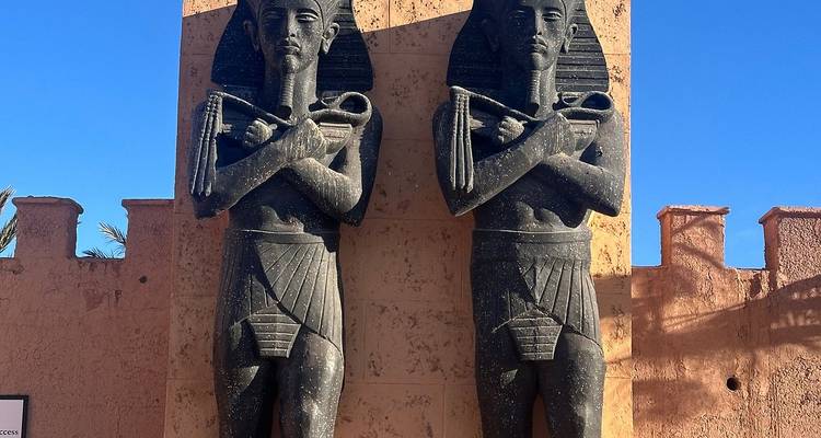 Twin pharaoh statues with crossed arms set against a bright blue sky at a film studio entrance.