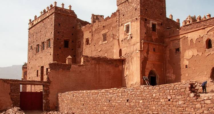 Kasbah en adobe patiné avec des tours crénelées et une porte en bois se détachant sur un ciel désertique pâle.