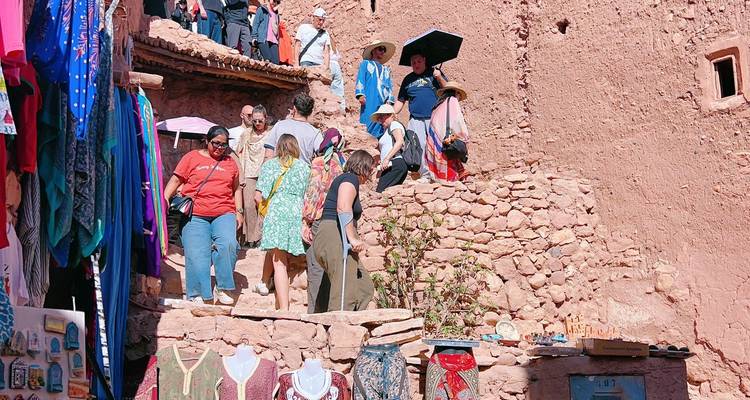 Tourists climb a narrow stone stairway lined with colorful textiles and souvenir stalls inside an adobe village.