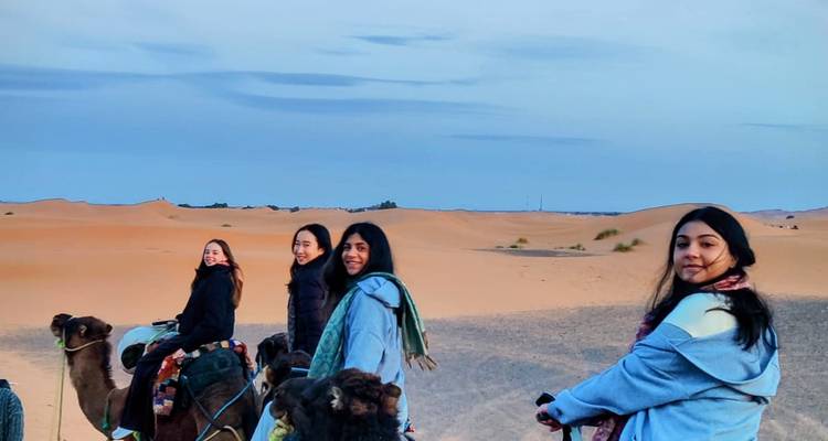 Four young women smile while riding camels across soft desert dunes beneath a pastel sky.