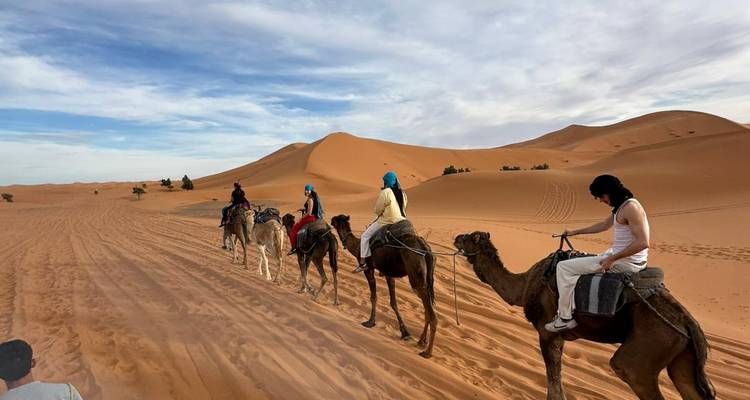 Riders in bright headscarves follow each other on camels along vehicle tracks in open dunes.