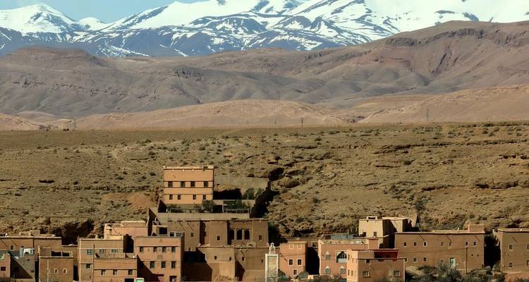 Earthen village sits on an arid plateau with snow-capped Atlas Mountains towering behind.