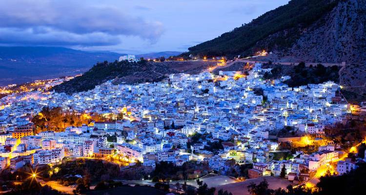 Paysage urbain crépusculaire montrant une ville à flanc de colline illuminée de lumières bleu-blanc sous des montagnes boisées.