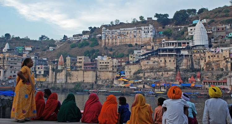 Kleurrijk geklede pelgrims zitten aan een rivier en kijken naar het tegenoverliggende oude stadslandschap