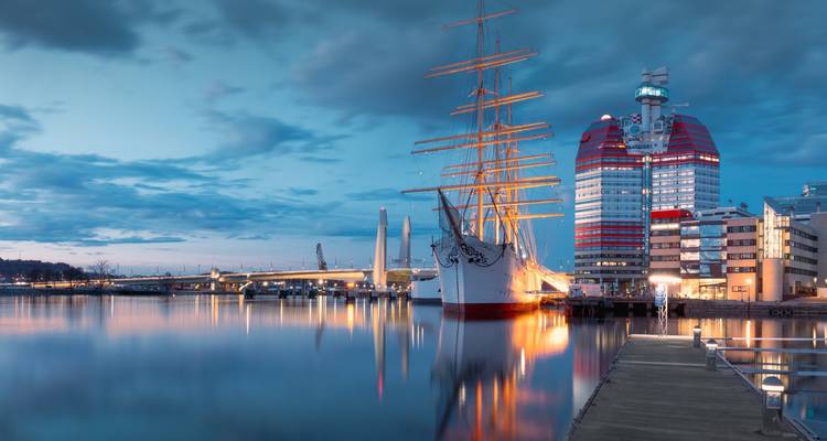 Dämmerungsansicht des Göteborger Hafens mit einem hohen weißen Segelschiff und dem rot-weißen Lipstick-Gebäude, die sich im ruhigen Wasser spiegeln