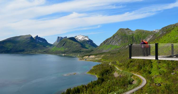 Zwei Besucher stehen auf einem auskragenden Aussichtspunkt hoch über einem Fjord und grünen Bergen