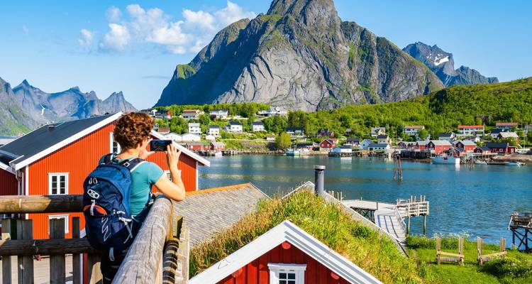 Rucksacktourist fotografiert rote Rorbu-Hütten und dramatischen Gipfel über einer blauen Bucht in Lofoten