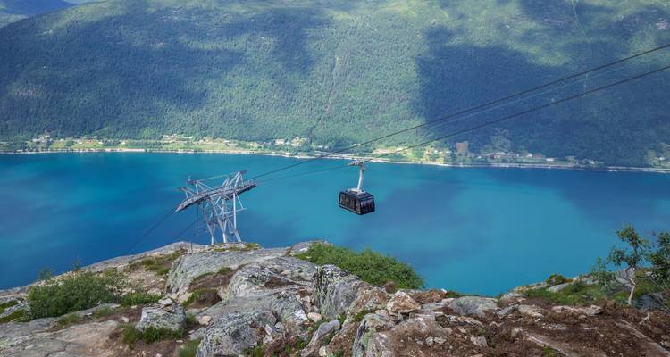 Seilbahn-Gondel, die hoch über einem tiefblauen Fjord gleitet, vor steilen grünen Bergen.