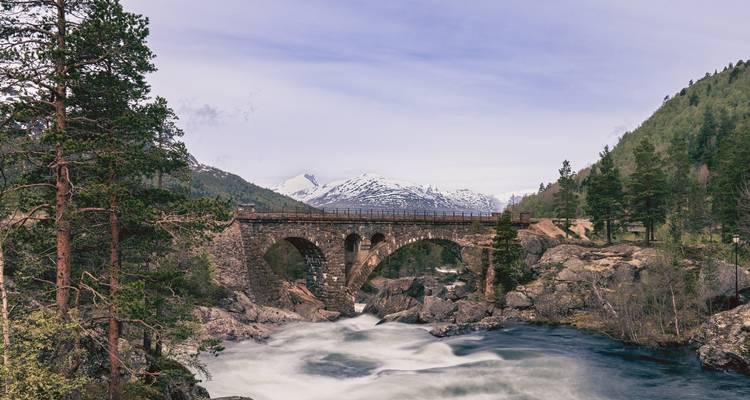Historische steinerne Bogenbrücke, die einen reißenden Gebirgsfluss überspannt, mit schneebedeckten Gipfeln dahinter.