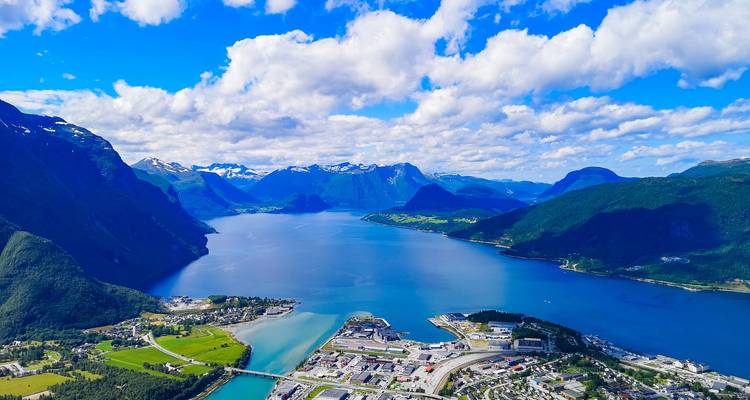 Blick aus der Vogelperspektive auf einen tiefblauen Fjord, umgeben von dramatischen Alpengipfeln und einer kleinen Stadt am Ufer.