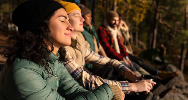 Eine entspannte Gruppe von Wanderern sitzt auf einem Felsen im herbstlichen Waldsonnenschein, eine Frau genießt die warmen Strahlen mit geschlossenen Augen.