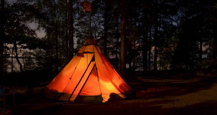 Ein oranges Tipi-Zelt leuchtet warm in einer dunklen Waldlichtung neben einem ruhigen See unter einem sternenklaren Himmel.