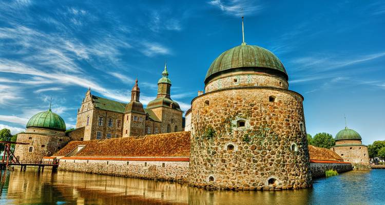 Das steinummauerte Schloss Vadstena mit runden Türmen spiegelt sich in seinem Wassergraben unter strahlend blauem Himmel.