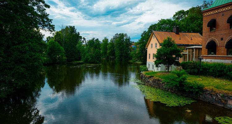 Ruhiger Fluss gesäumt von Bäumen, Seerosen und historischen Häusern unter stimmungsvollem Himmel.